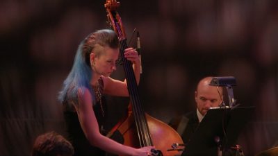 Woman with blue hair wearing a ball gown, playing upright bass, next to a drummer.