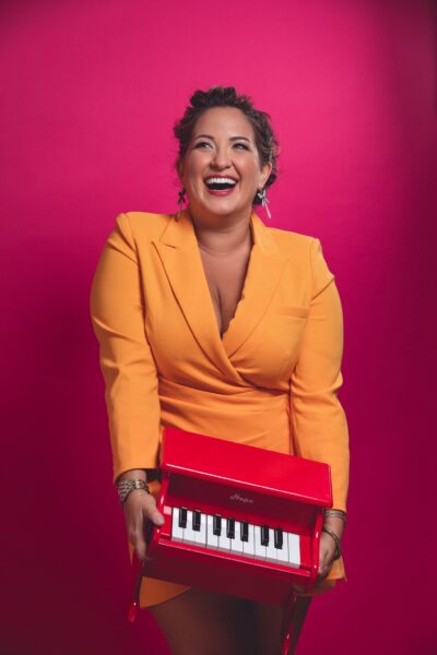 Woman smiling holding a small red piano wearing bright colors.