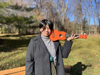 Julienne K. Tsang holds an orange ukulele like a violin even though they've never played violin