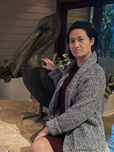 Brandy, a woman of Asian and European descent, is pictured wearing a large gray cardigan and resting her hand on a sculpture of a pelican.